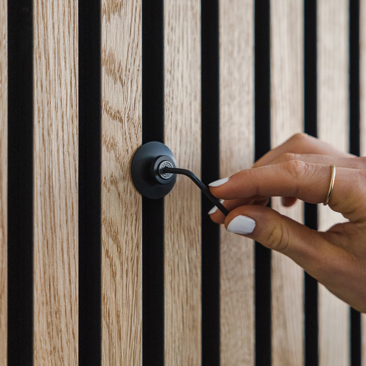 A hand with white nail polish turns a Dupps | Heavy black aluminium hook by WoodUpp on a vertical Akupanel acoustic wood panel.